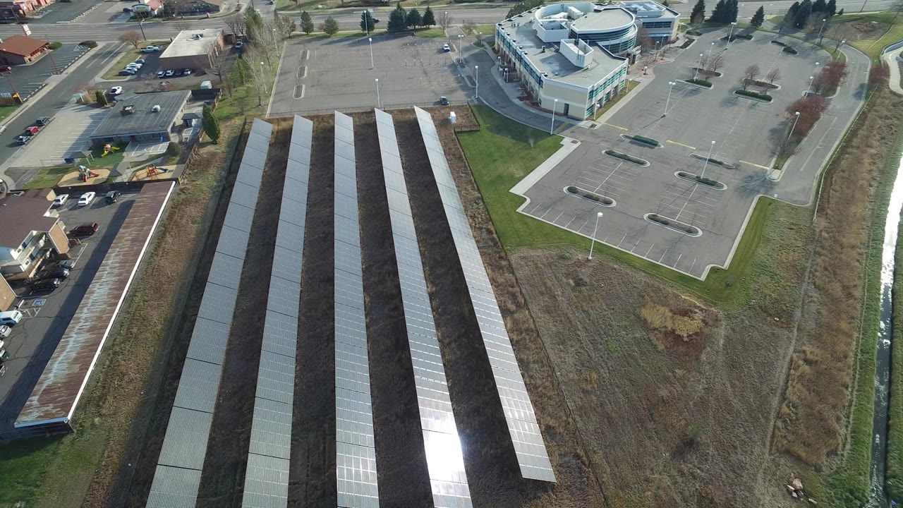Aerial frame in Westminster, Colorado with parallel solar panel rows dominating the lower half, a bright reflection on one row, an empty parking lot to the right, and no visible people.