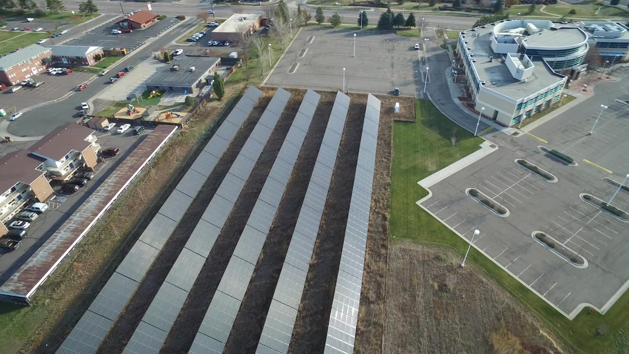 Aerial view of solar panel rows beside parking lots and low buildings.