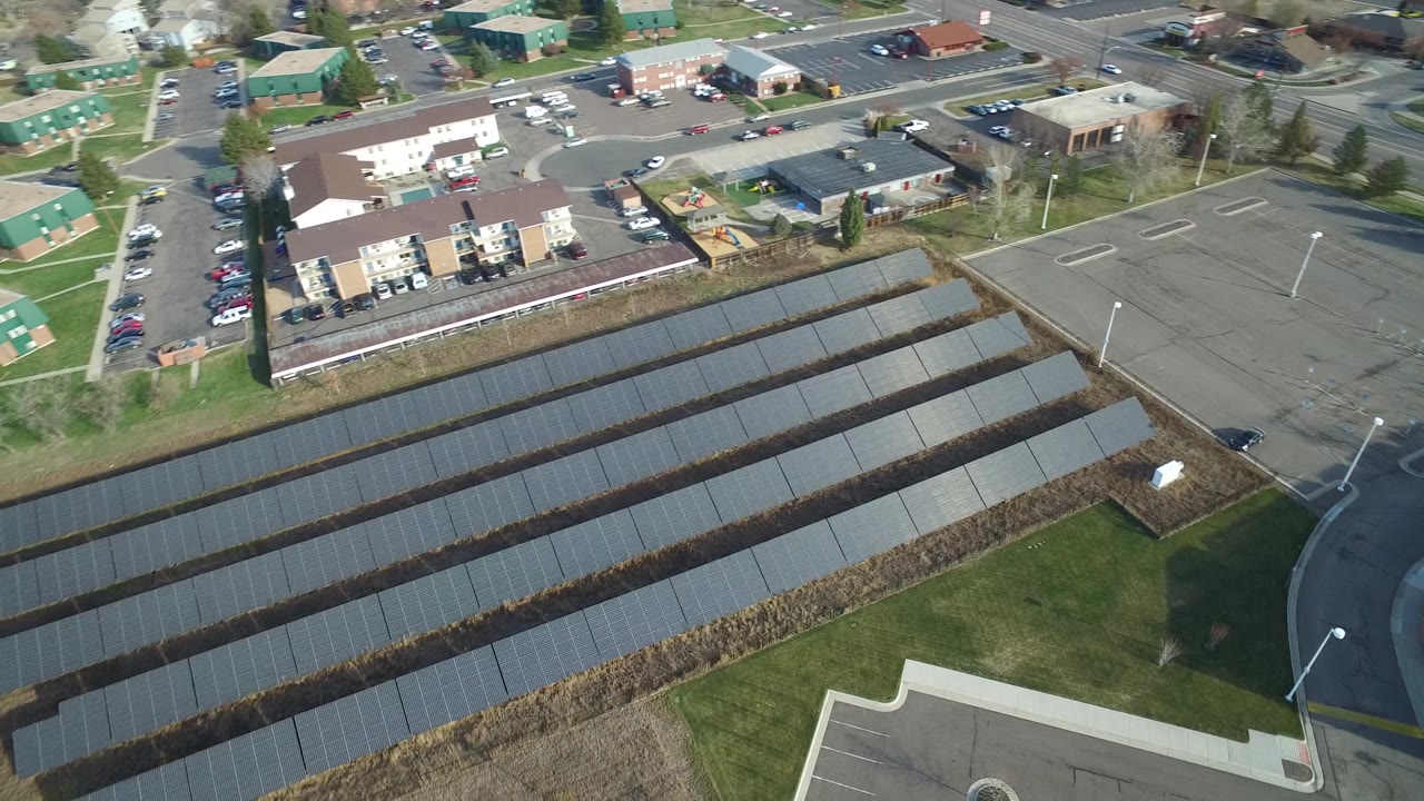 High aerial frame in Westminster, Colorado with the solar panel rows centered, a curving road at right, apartment blocks beyond, and no visible people.
