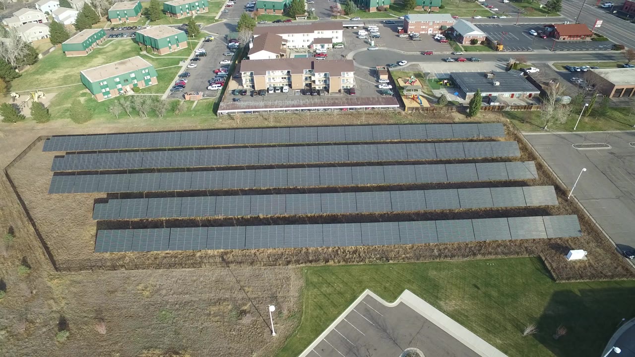 High aerial view over Westminster, Colorado with long rows of dark solar panels in the foreground, apartment-style buildings and parking lots beyond, and no visible people.
