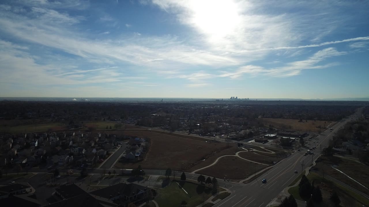 Wide aerial view over Westminster, Colorado suburbs with roads, open fields, a distant skyline, strong sun glare, and no visible people.