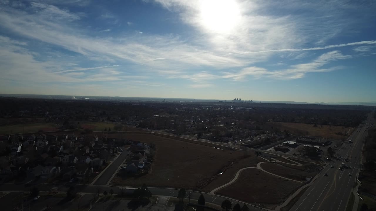 This Westminster, Colorado aerial frame keeps the sun high above a dark residential foreground, brown open land, a straight road, and small distant buildings on the horizon, with no visible people.
