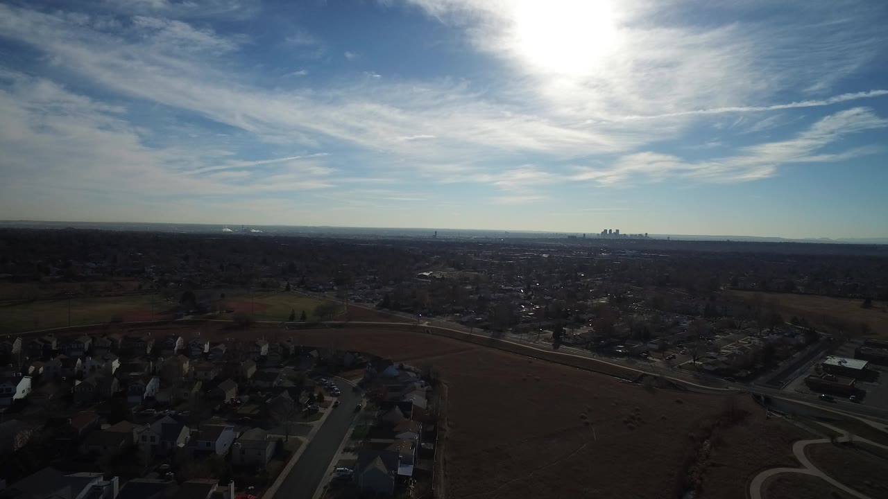 High drone view across Westminster, Colorado residential streets and brown fields, with the sun near the top of frame and no visible people.