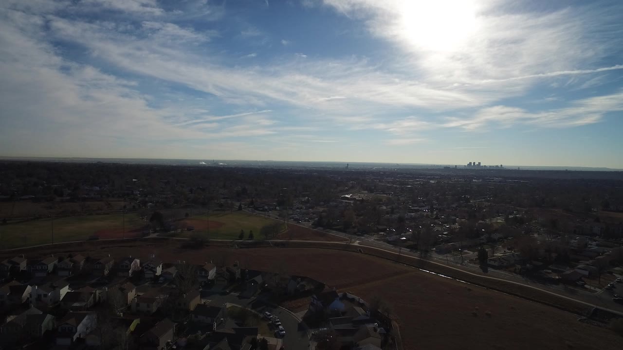High aerial frame over Westminster, Colorado with dark suburban rooftops in the foreground, open fields beyond, a distant skyline on the horizon, and no visible people.