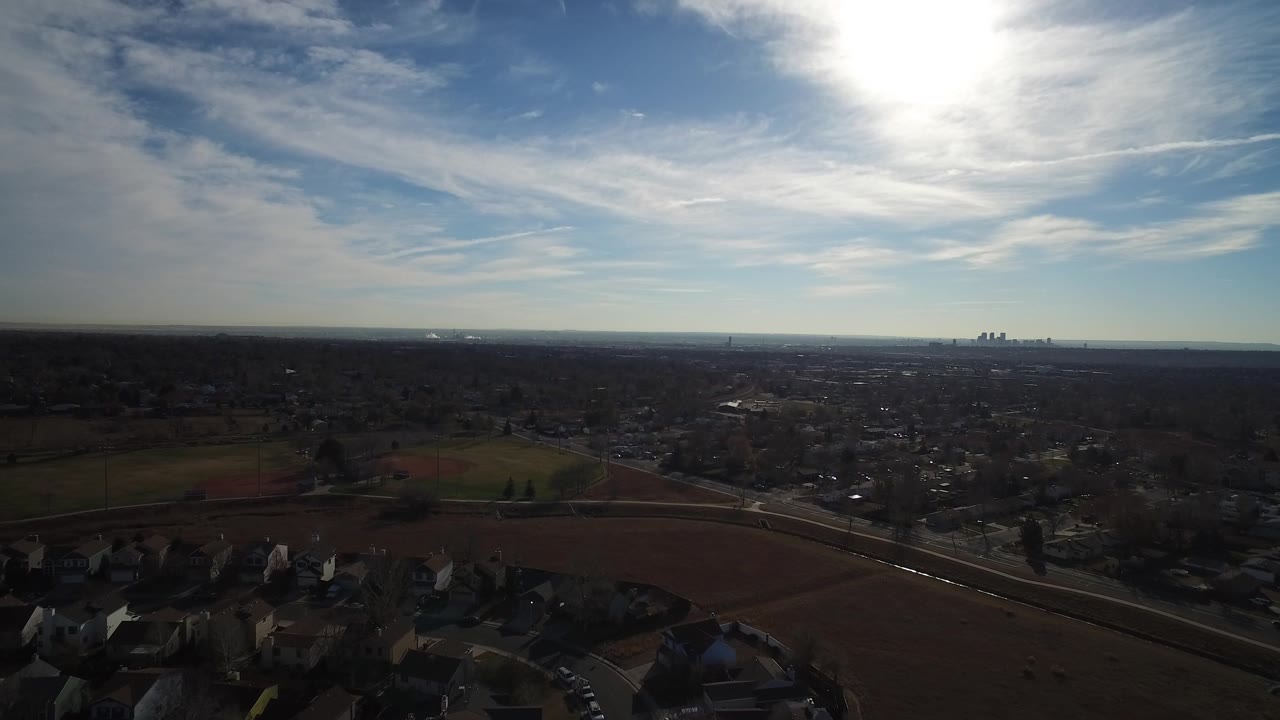 Wide drone-like view across rooftops and fields toward a low skyline under thin clouds.