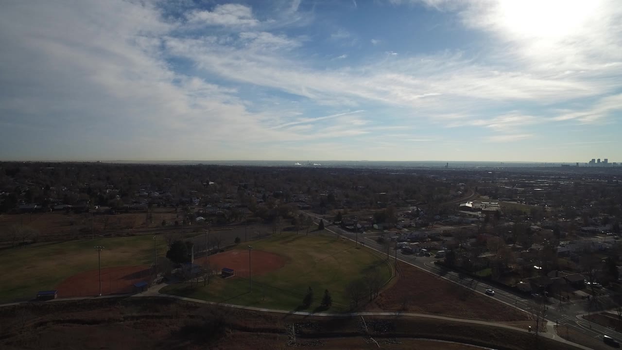 Aerial view over baseball fields, roads, neighborhoods, and a distant skyline under bright sun.