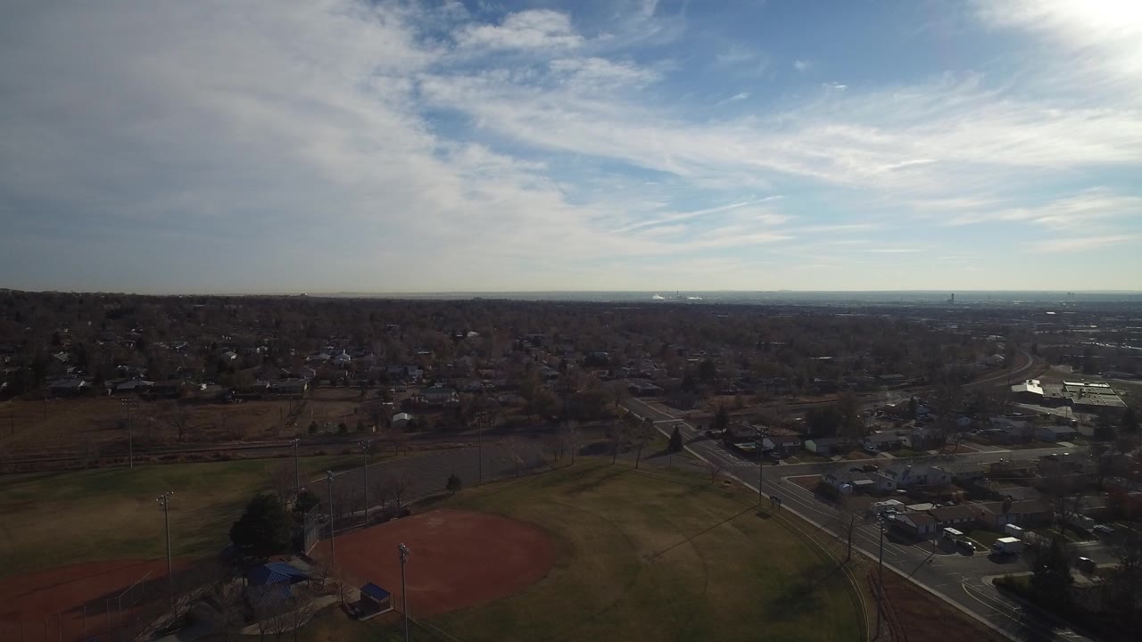 Wide aerial frame over Westminster, Colorado with ball fields in the foreground, roads and parking areas below, neighborhoods stretching to the horizon, and no visible people.