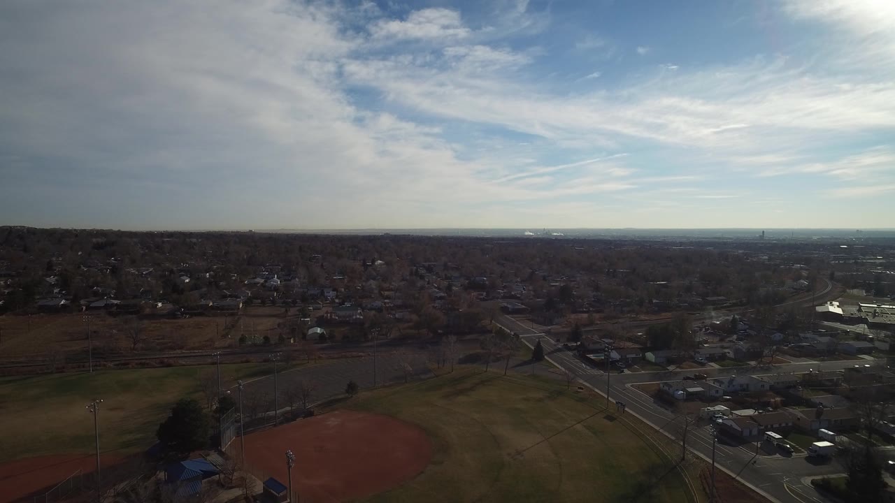 Drone-height view of ball fields, streets, parking lots, and neighborhoods beneath clouds.