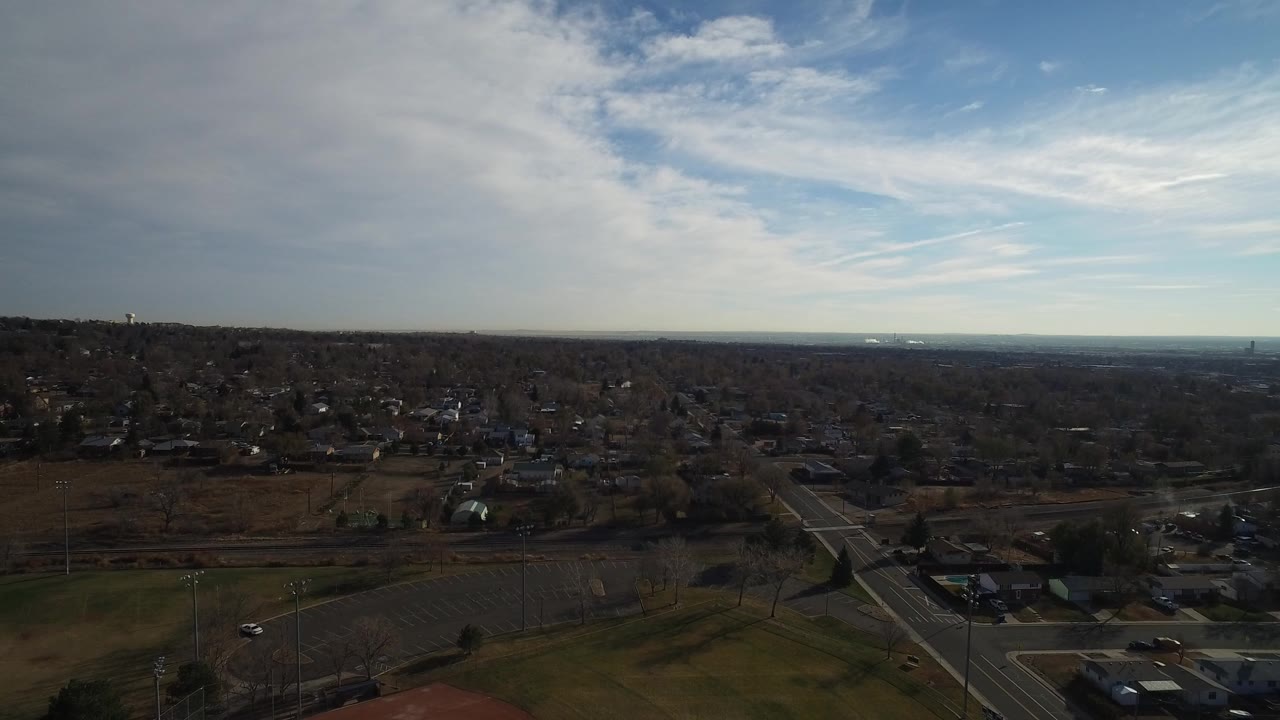 Wide aerial frame over Westminster, Colorado, showing a green field, roads, low neighborhoods, distant horizon, and no visible people.