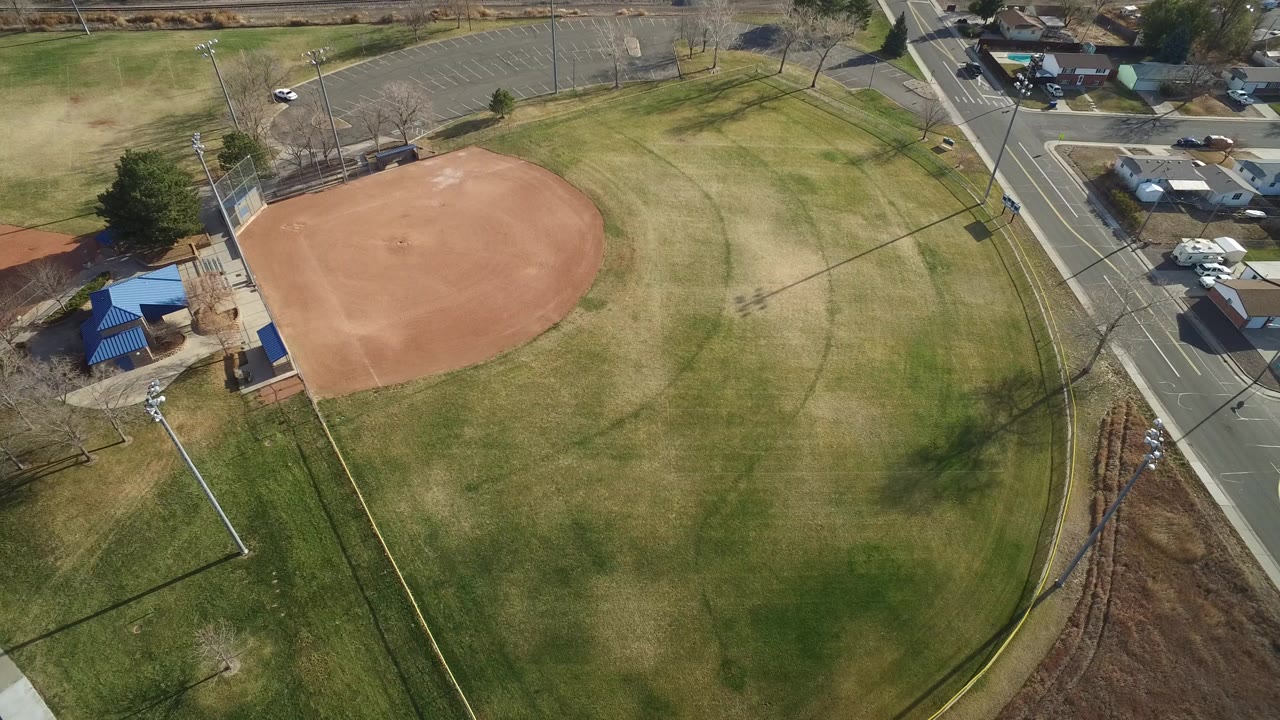 Aerial view of an empty baseball diamond beside grass, roads, and a blue-roofed park structure.