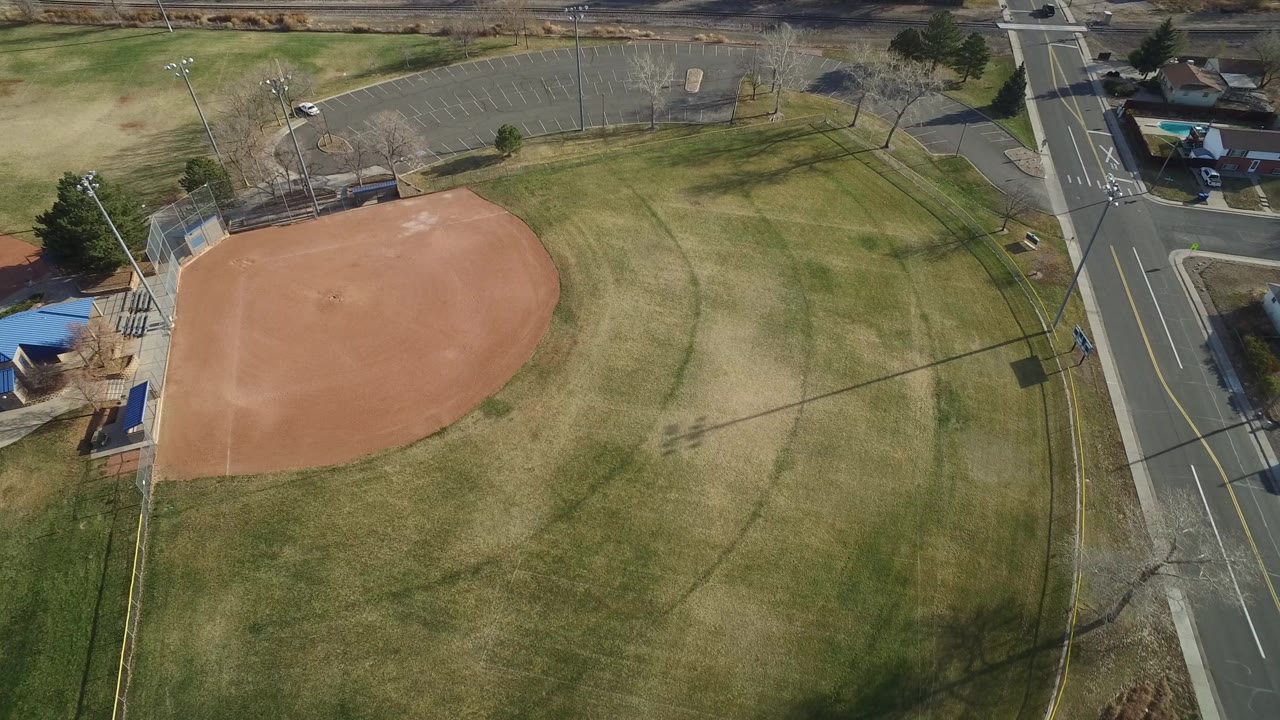 High aerial view of an empty baseball field in Westminster, Colorado, with the tan infield at left, open grass across the center, a road on the right, and no visible people.