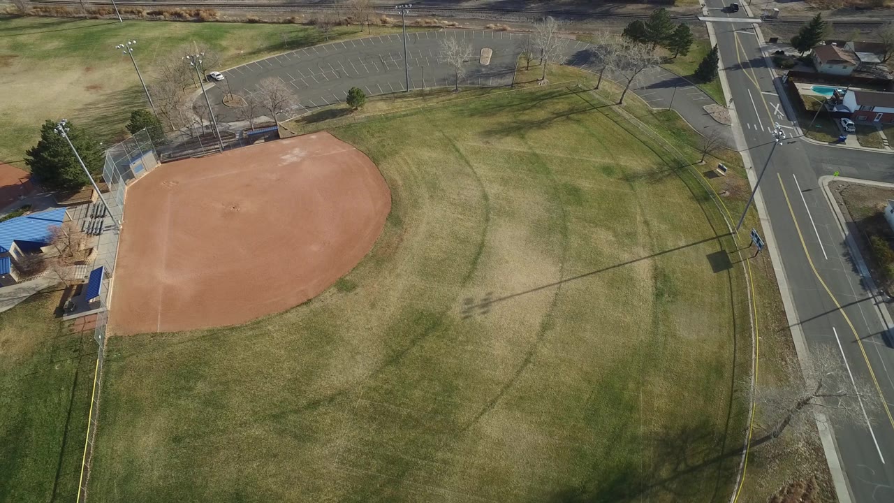 Aerial frame over Westminster, Colorado showing the empty ballfield slightly closer in the grid, with the infield on the left, grass outfield in the middle, roadside curb at right, and no visible people.
