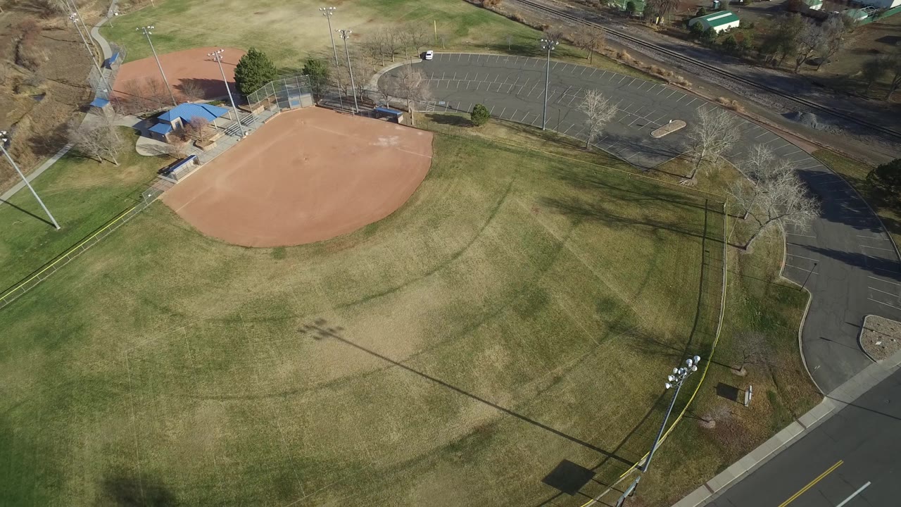 Aerial frame over the Westminster ballfield, showing the infield near the top, broad grass outfield, light-pole shadows, a curved roadside, and no recognizable people visible.