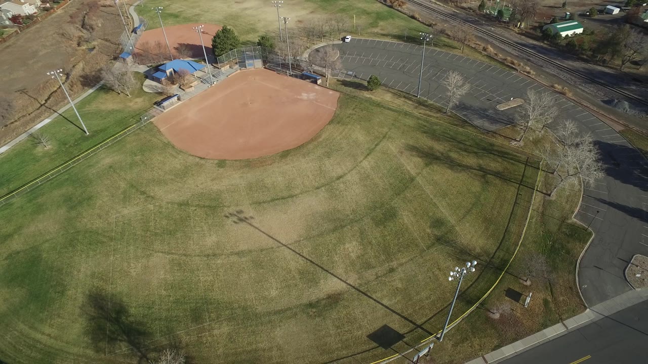 High drone view of the Westminster baseball field with the infield across the upper center, a dark parking lot to the right, rail tracks behind it, and no recognizable people visible.