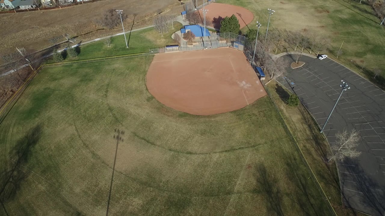 Drone-style overhead view in Westminster, Colorado with the baseball diamond high in frame, a dark paved parking lot to the right, grass fields below, and no visible people.