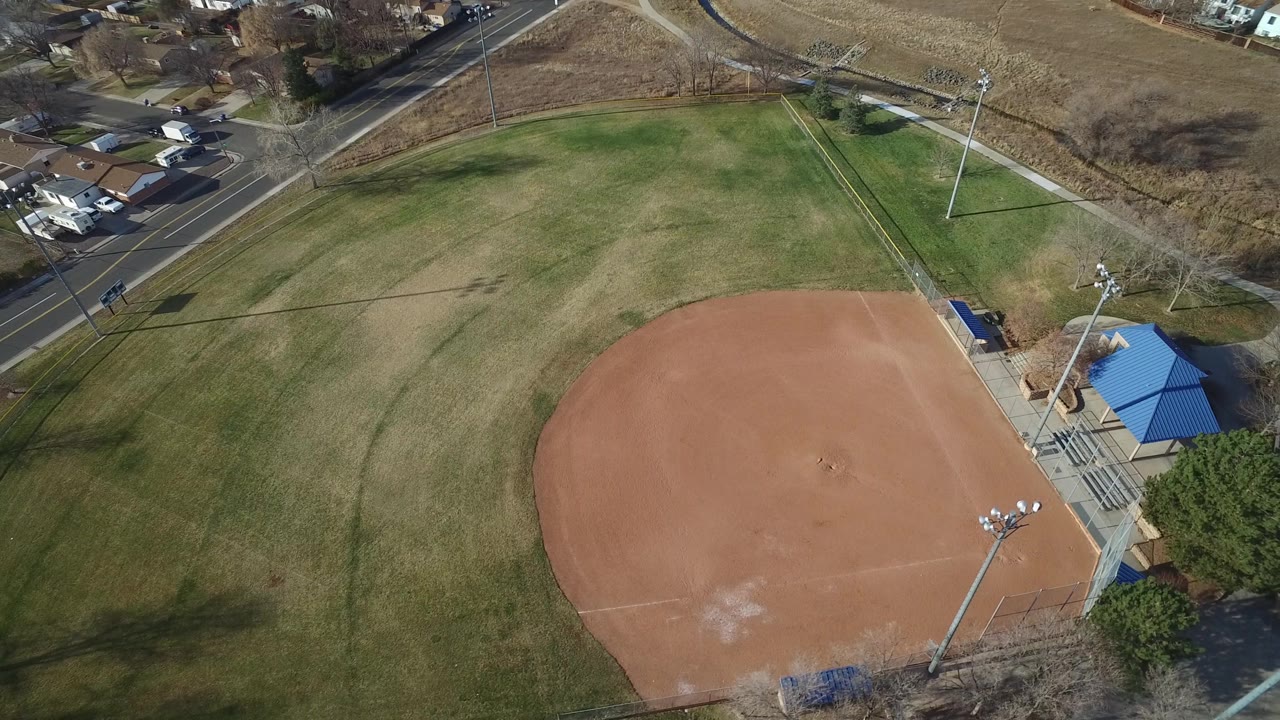 The baseball diamond remains centered from above in Westminster, Colorado, with the blue-roofed structure on the right edge, residential homes beyond the road, and no visible people.
