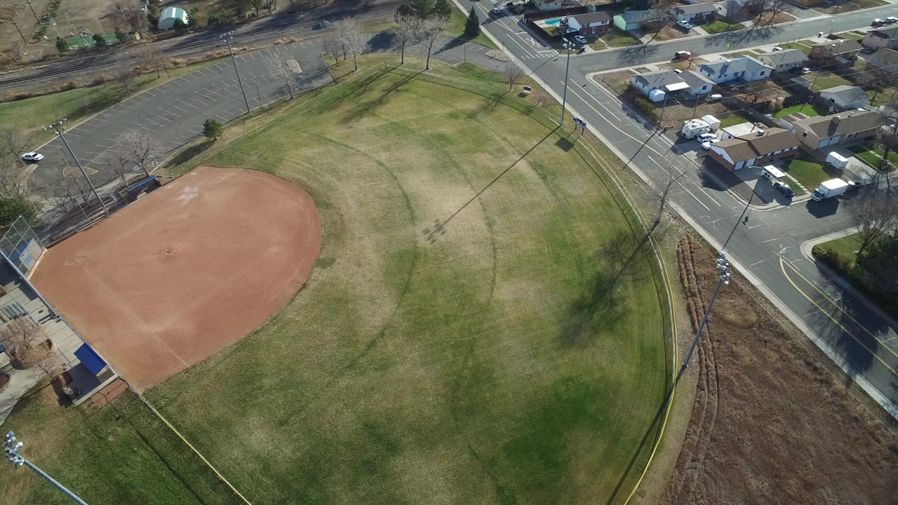 Aerial view in Westminster, Colorado, looking down on a red dirt ball field, surrounding grass, roads, parked vehicles, and small buildings, with no visible people.