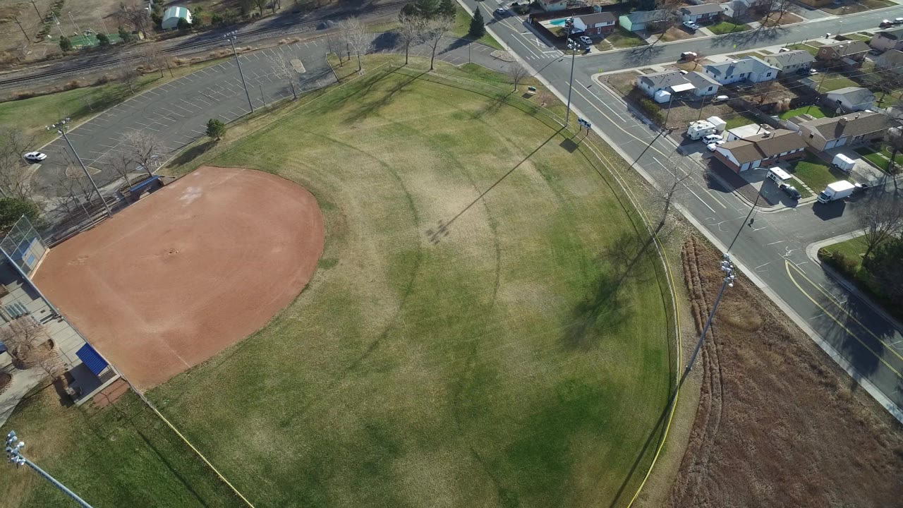 Overhead Westminster, Colorado frame with the ball field shifted toward the lower left, a road intersection above it, parked vehicles near small buildings, and no visible people.