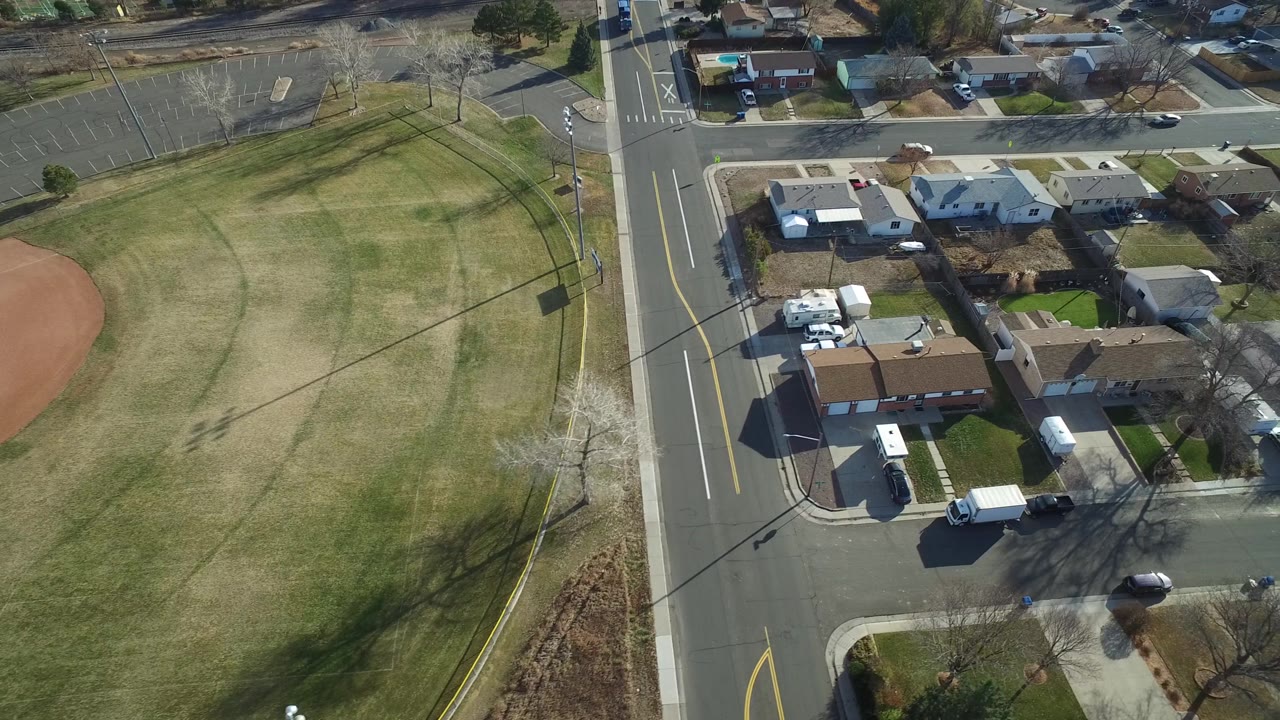 Aerial view in Westminster, Colorado with the grass field on the left, a curved road near center, residential roofs and driveways on the right, and no visible people.