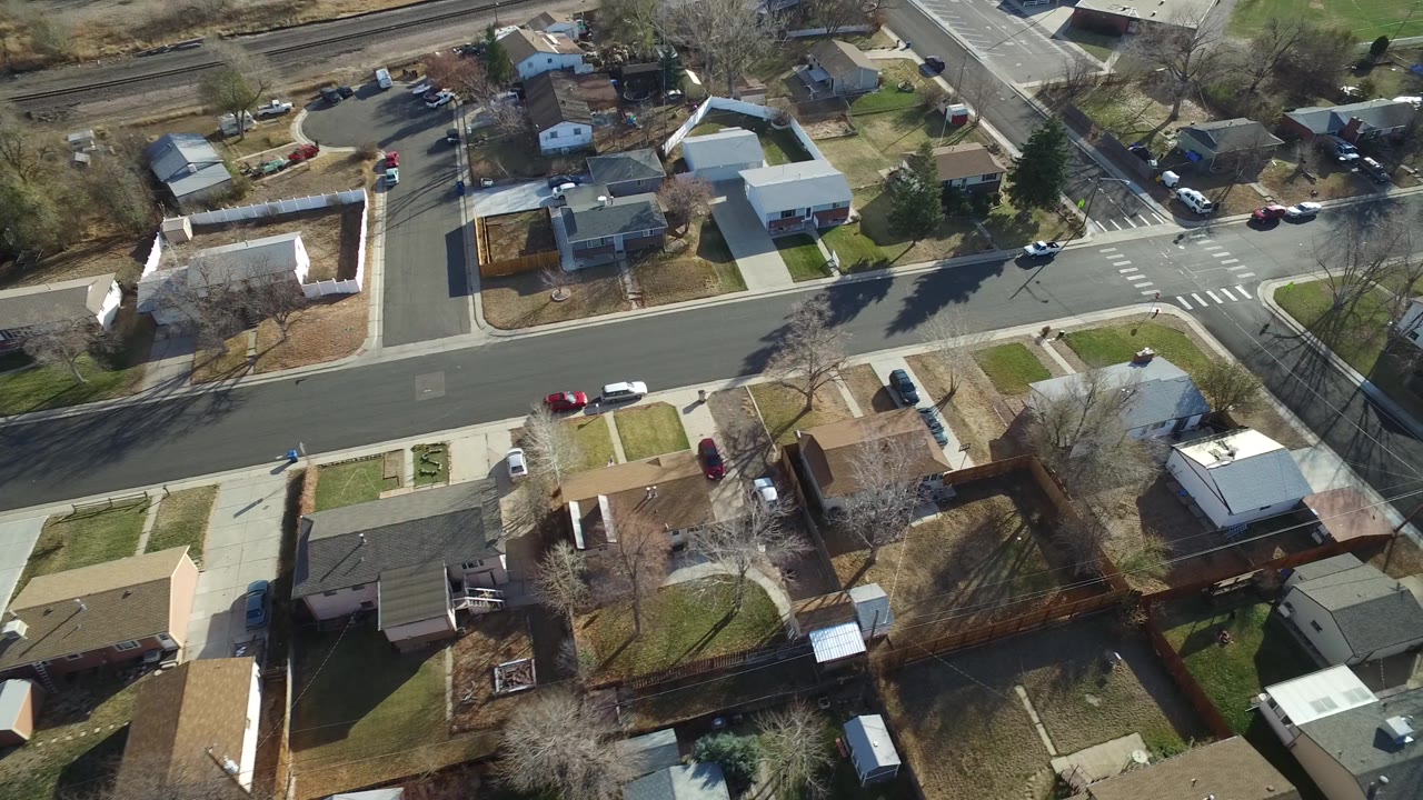 Drone-view frame in Westminster, Colorado with rooftops, driveways, a crosswalk-marked intersection, parked cars along the curb, rail tracks in the background, and no visible people.