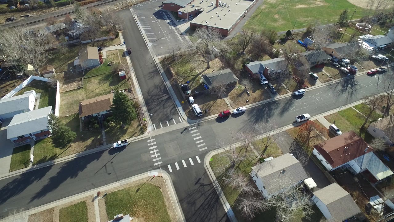Aerial view of a residential intersection with houses, parked cars, crosswalks, and a nearby field.