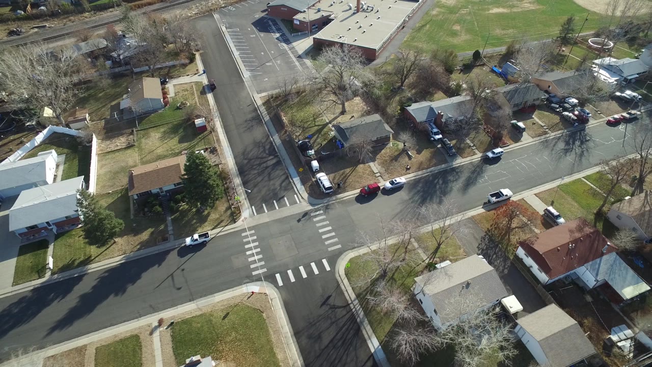 Oblique drone view in Westminster, Colorado with a neighborhood road in the foreground, several parked vehicles, houses with yards, a large parking lot, and a green field beyond, with no clearly visible people.