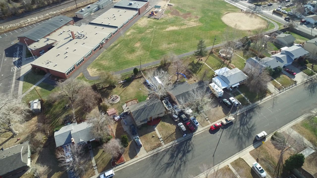 Aerial frame over Westminster, Colorado with the school roof at left, open grass and a baseball diamond at top, houses along the street, parked vehicles, and no clearly visible people.