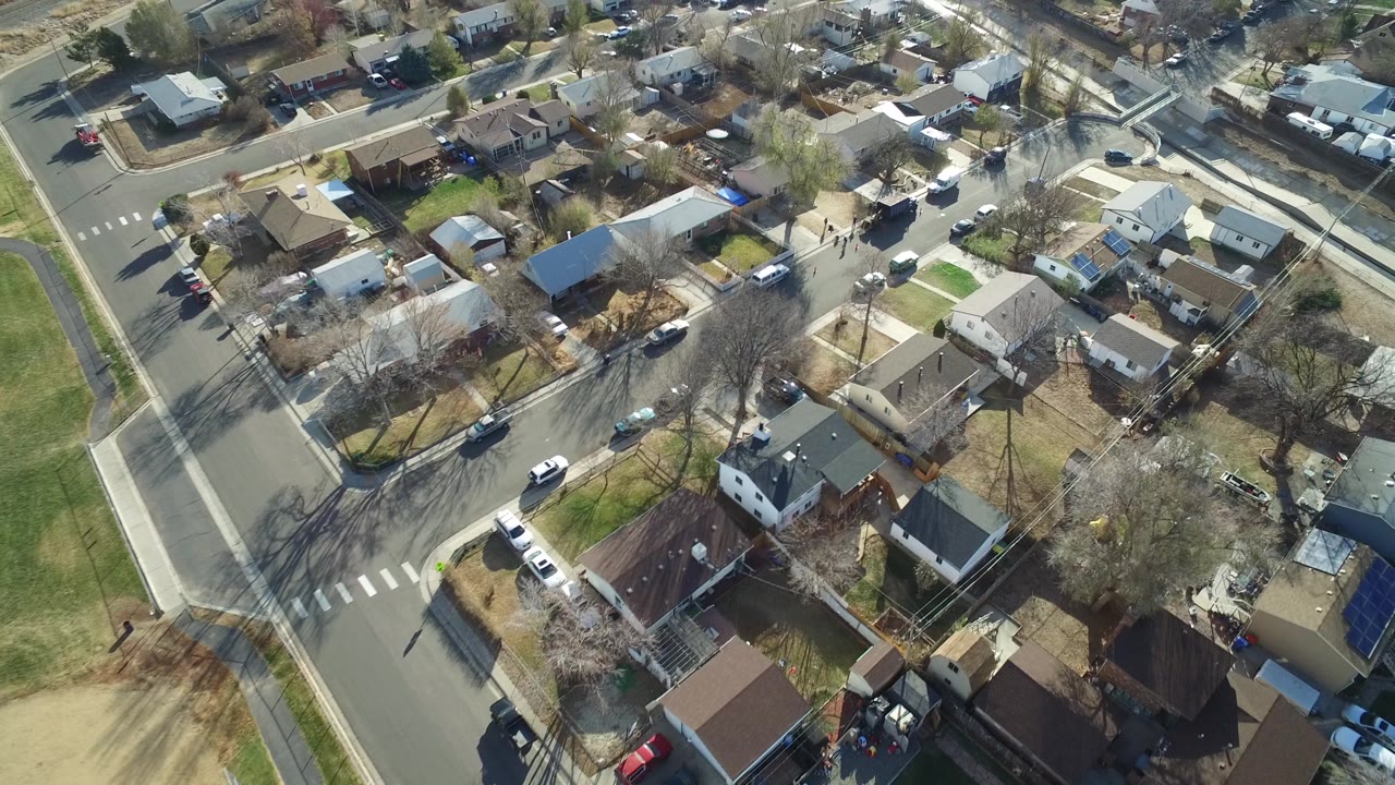 Aerial frame over Westminster, Colorado homes near a street corner, showing rooftops, yards, cars along curbs, bare tree canopies, and no visible people.