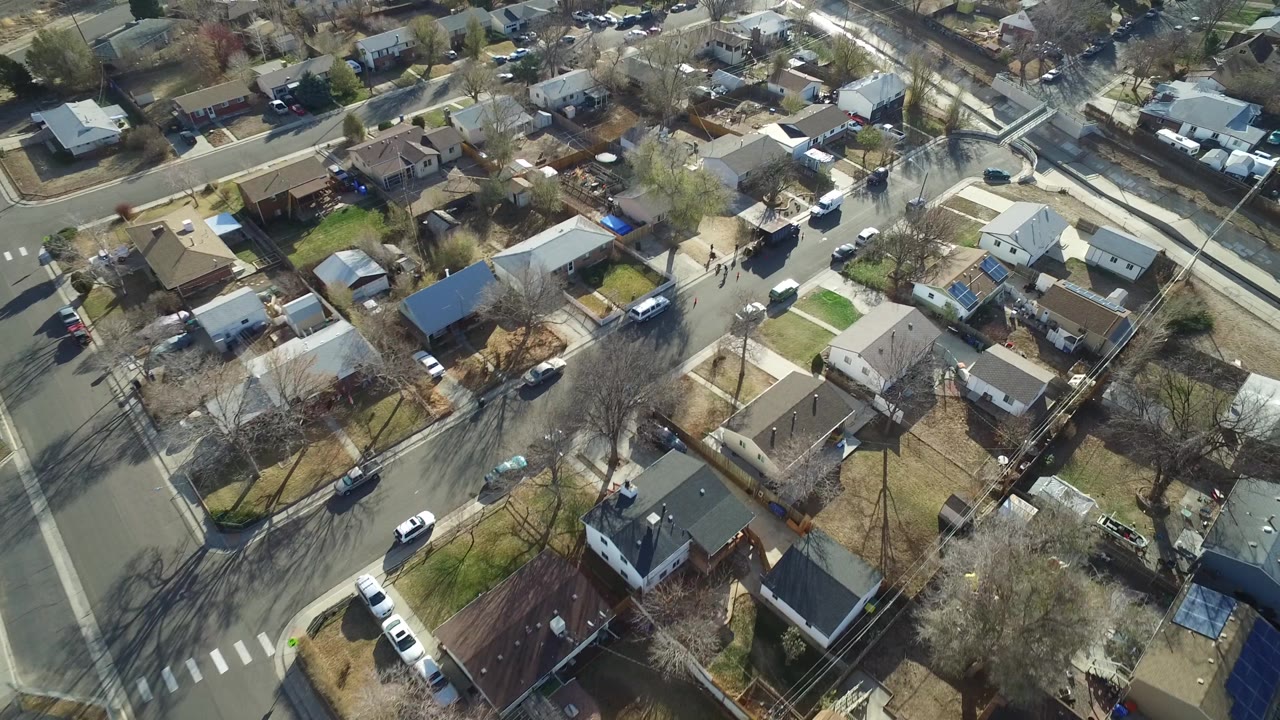 Aerial neighborhood view with houses, lawns, roads, and a long pale path.