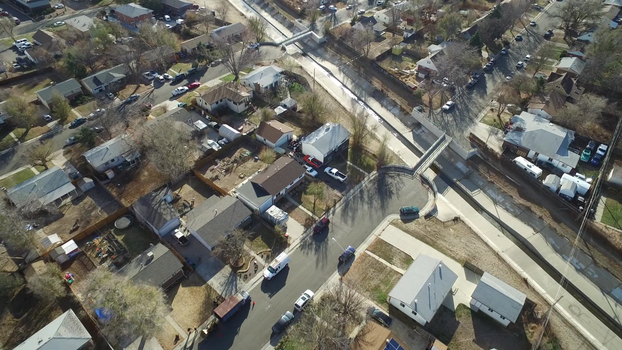 Aerial view over a Westminster, Colorado neighborhood with rooftops, fenced yards, parked cars, bare trees, and a pale canal running diagonally through the frame, with no visible people.