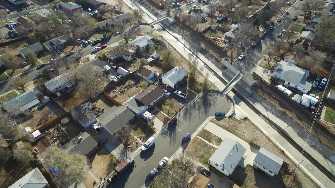 High overhead frame of Westminster, Colorado homes, driveways, parked vehicles, leafless trees, and a light-colored canal path crossing beside a curved residential street, with no visible people.
