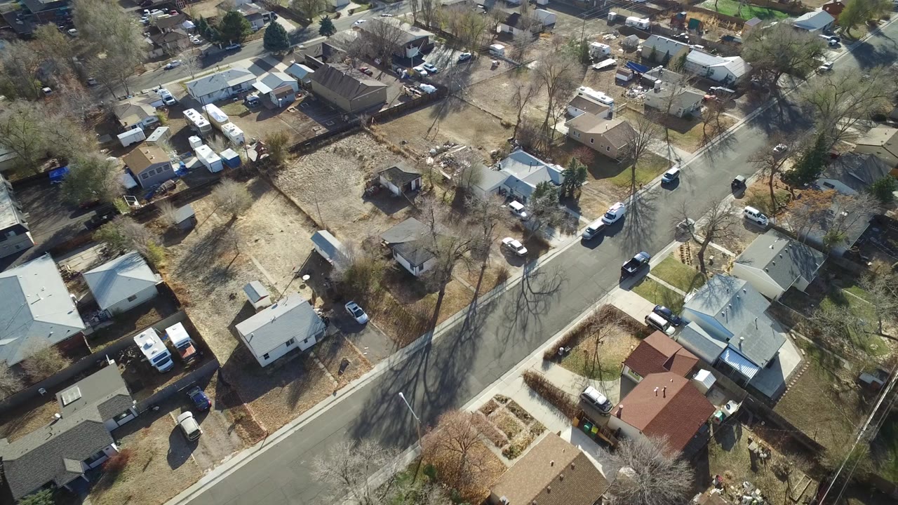 Aerial frame over Westminster, Colorado showing a curving residential road, detached homes, bare trees casting shadows, and several parked vehicles, with no visible people.