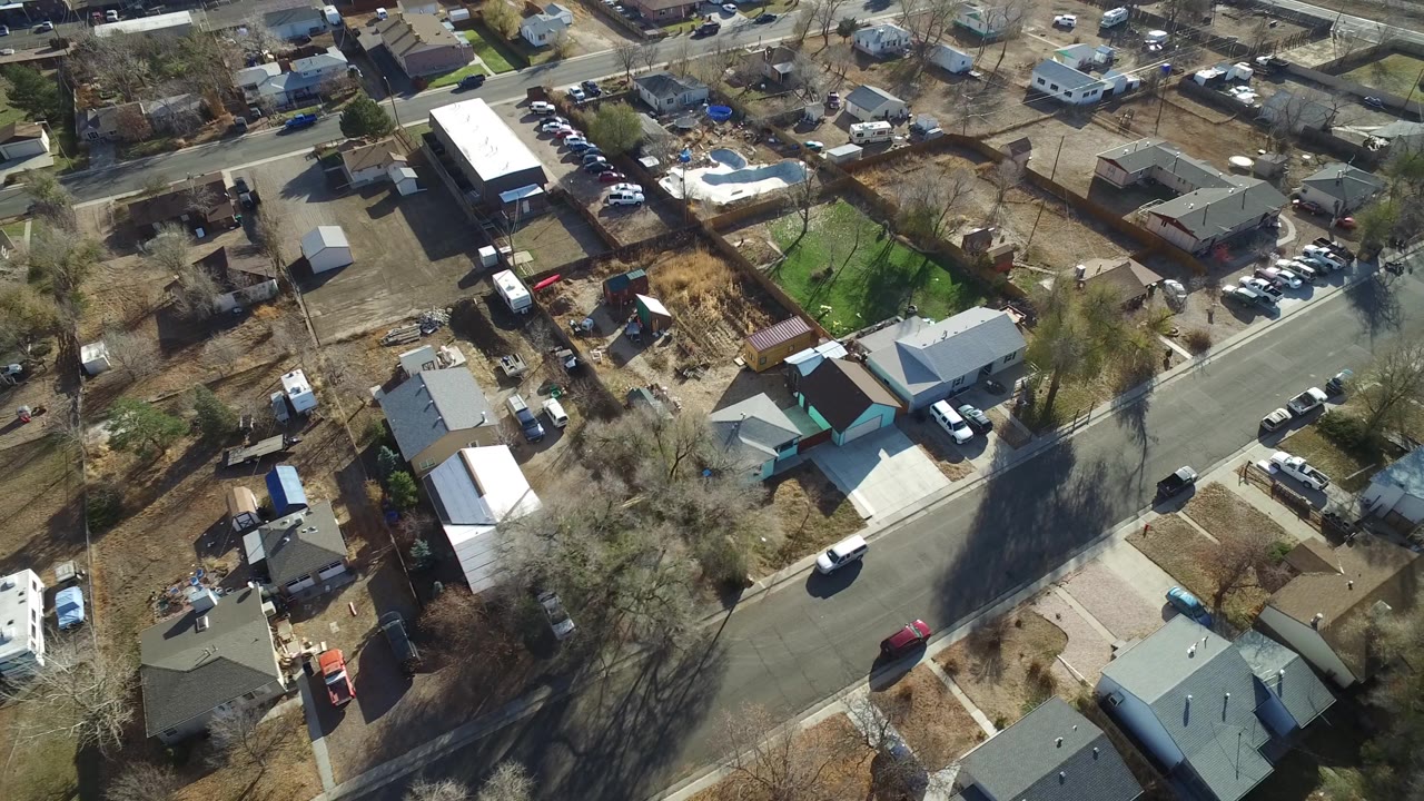 High overhead frame in Westminster, Colorado showing a diagonal neighborhood road, clustered rooftops, fenced backyards, parked cars, and no clearly visible people.