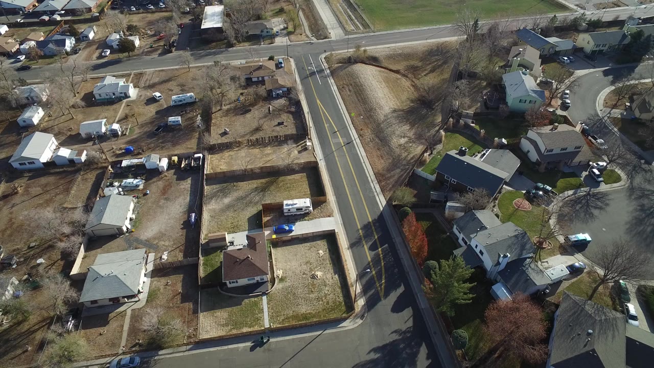 High aerial view over Westminster, Colorado houses beside a two-lane road and a cul-de-sac, with fenced yards, parked vehicles, leafless trees, and no visible people.