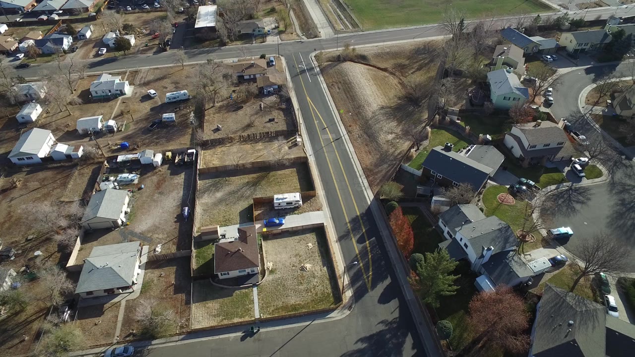Aerial frame in Westminster, Colorado showing a road intersection beside detached homes, bare trees, driveways, parked cars, and open tan yards with no visible people.