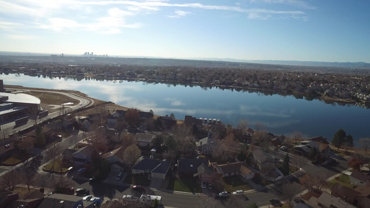 High aerial frame over Westminster, Colorado homes and curving roads beside a broad water body, with a distant skyline on the horizon and no visible people.