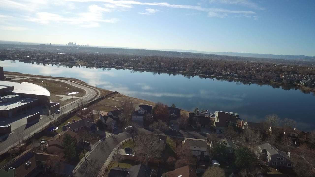 High aerial frame in Westminster, Colorado looking over a broad reflective water surface toward tree-lined houses, distant buildings, pale hills, and no visible people.