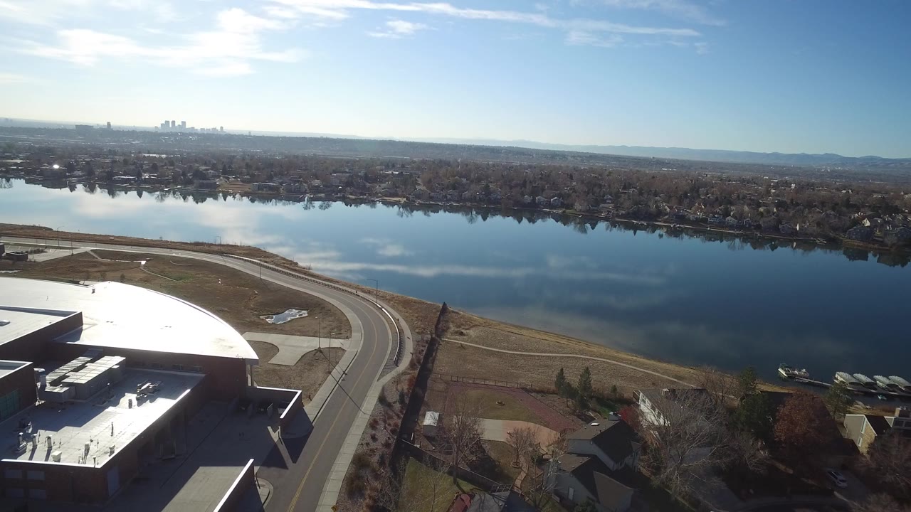 Aerial frame in Westminster, Colorado showing the corner of a large white-roofed warehouse, a curved road, open grass, a reflective lake, distant homes, and no visible people.