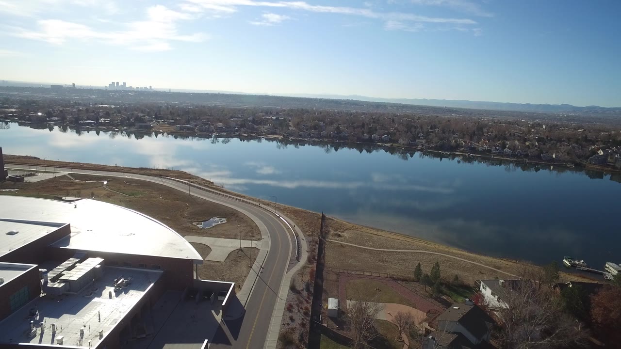 Aerial frame in Westminster, Colorado with the bright warehouse roof crossing the lower frame, a road and shoreline below, a wide lake, distant tree-lined homes, and no visible people.