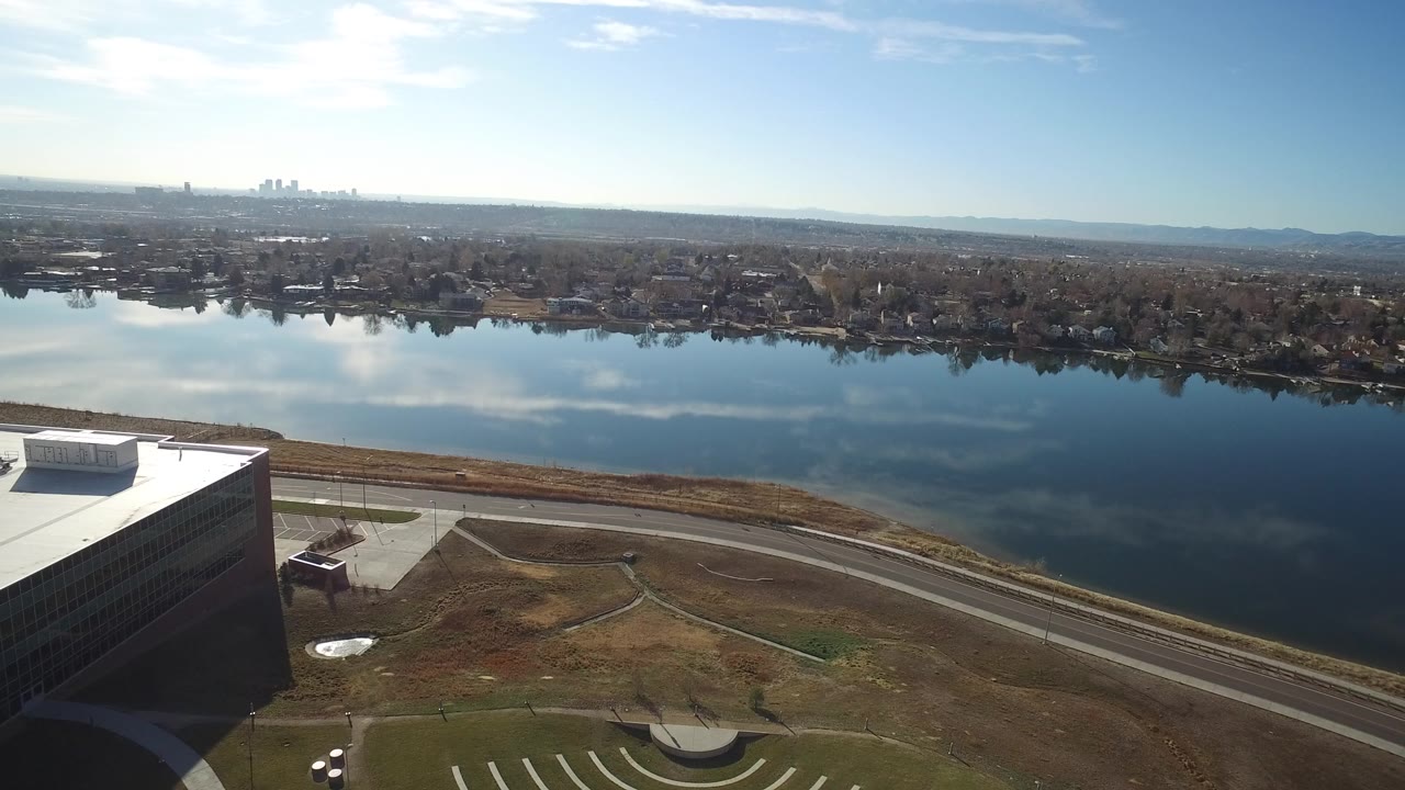 Aerial frame over Westminster, Colorado showing a white-roofed building at lower left, a calm lake with cloud reflections, shoreline homes, distant urban buildings, and no visible people.