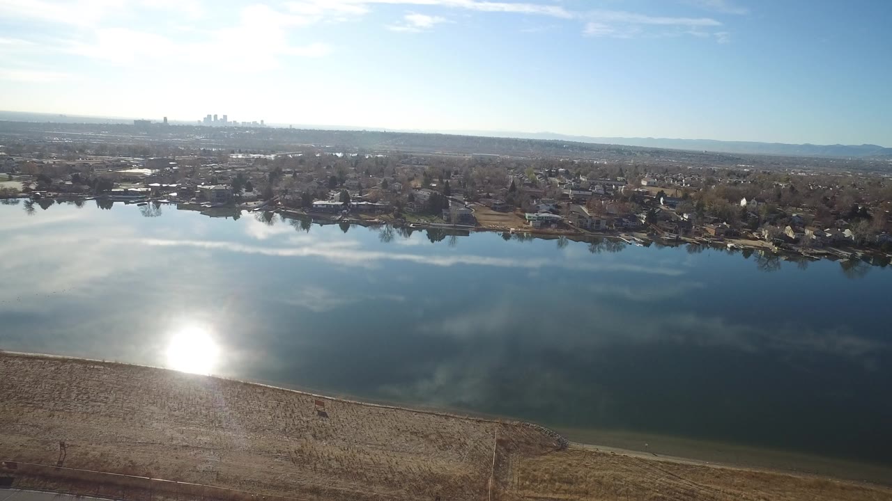 Aerial view of a lake with bright sun reflection and suburban homes beyond the water.