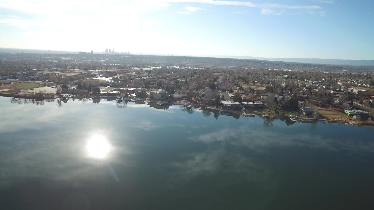 Aerial view over reflective water toward shoreline homes and a distant skyline.