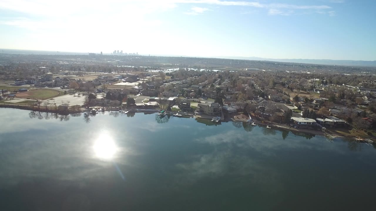 Aerial view in Westminster, Colorado, looking across a lake with strong sun glare on the water, shoreline houses, open fields, and no visible people.