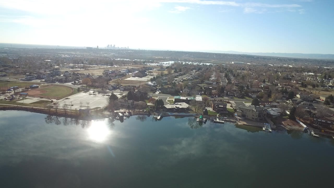 Aerial view over water toward homes, sports fields, and a distant skyline.