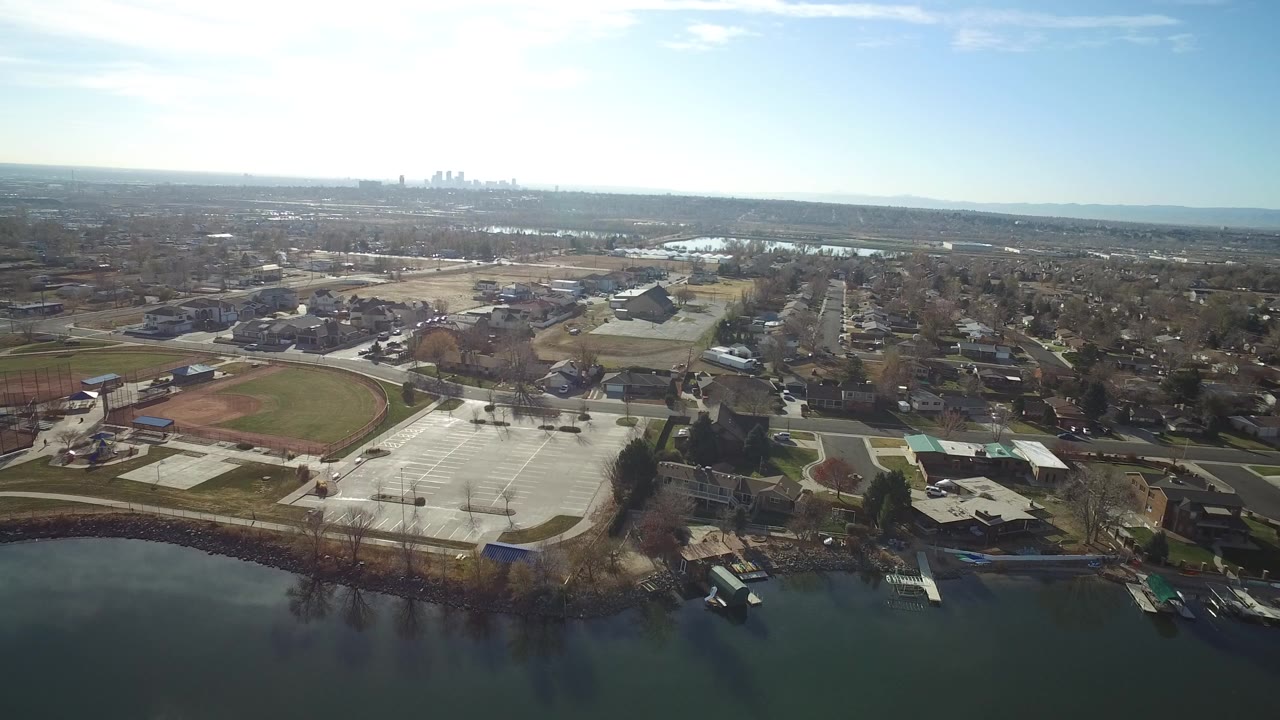 Aerial view over water toward sports fields, parking lots, homes, and a distant skyline.