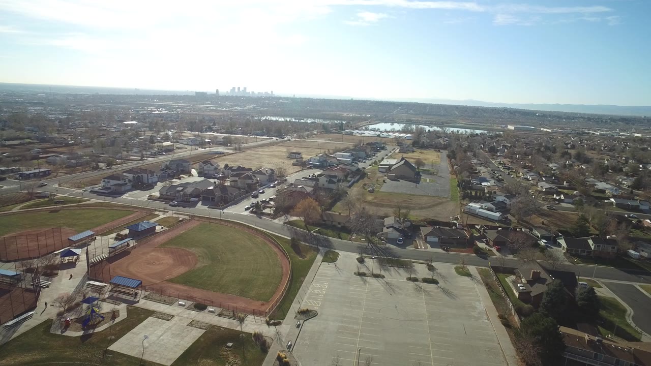 Aerial view in Westminster, Colorado with the baseball field more centered, fenced courts on the left, parking pavement below, homes beyond, and no visible people.