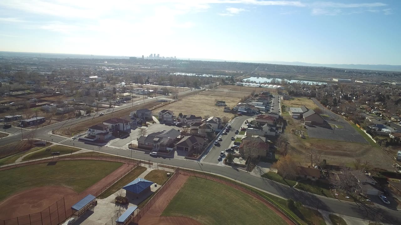 Opening aerial frame over Westminster, Colorado, with homes and roads below, a ballfield corner at the bottom, distant water near the horizon, and no visible people.
