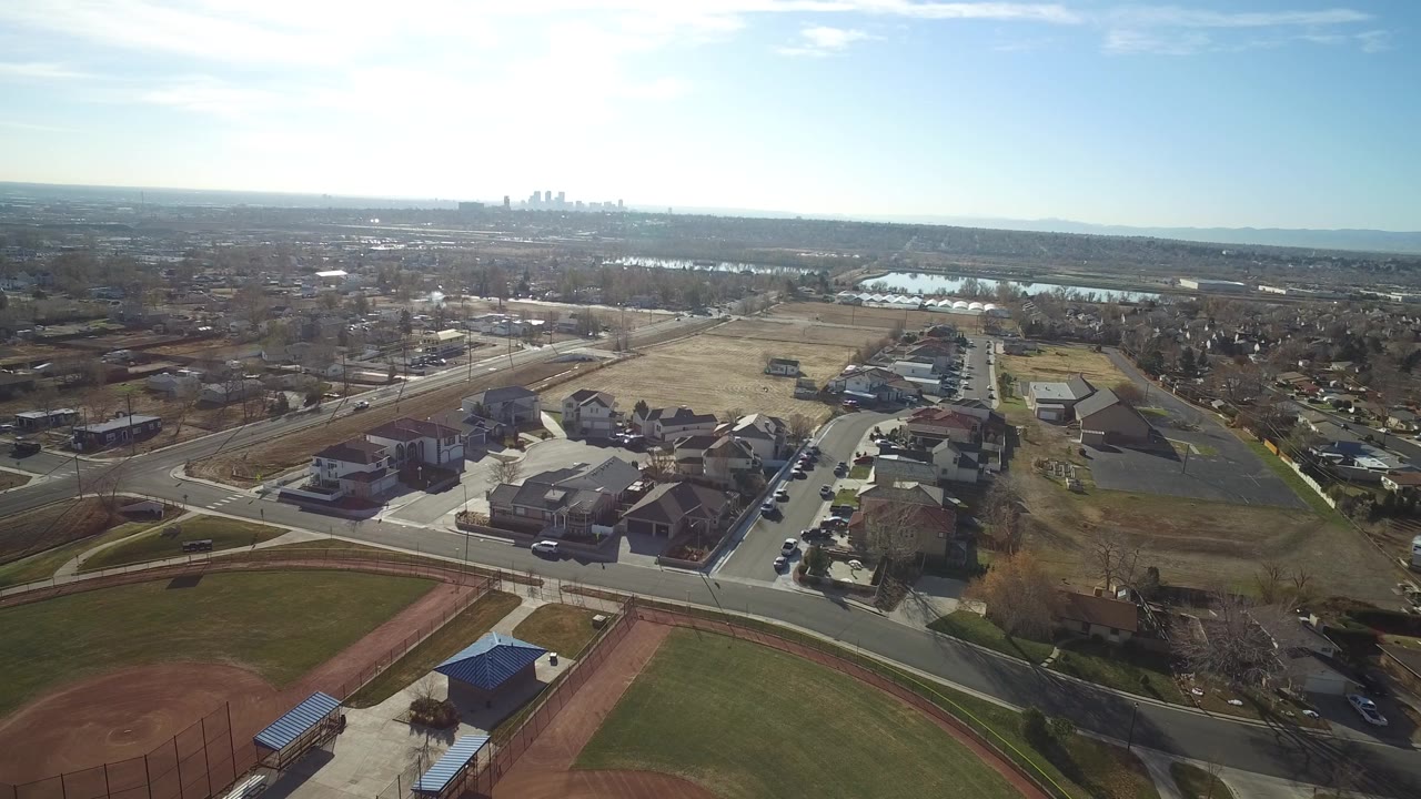 Aerial view over Westminster, Colorado, with rows of houses, a road intersection, pale open ground, distant lakes, a far skyline, and no visible people.