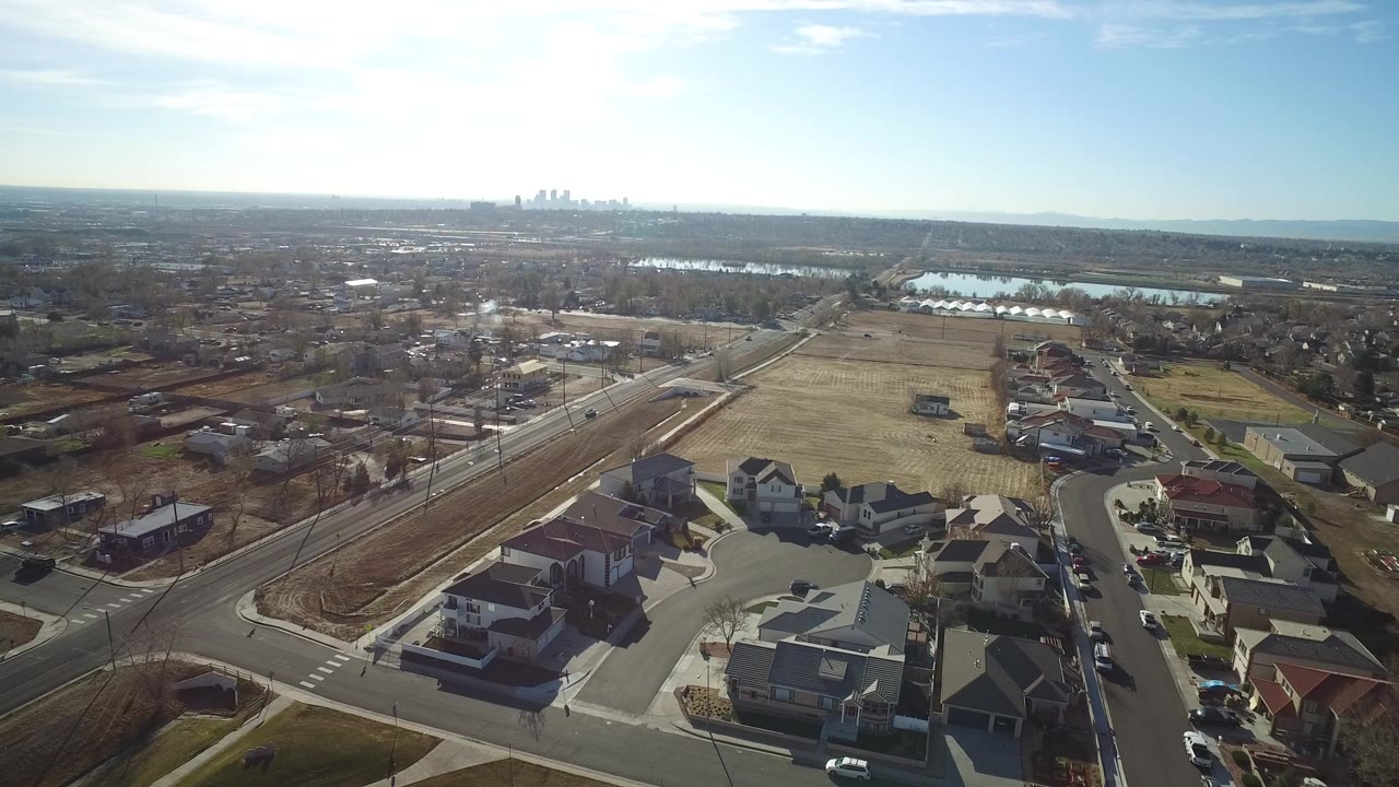Opening aerial frame over Westminster, Colorado, with private homes and curved streets in the foreground, open fields and water beyond, a hazy skyline on the horizon, and no visible people.