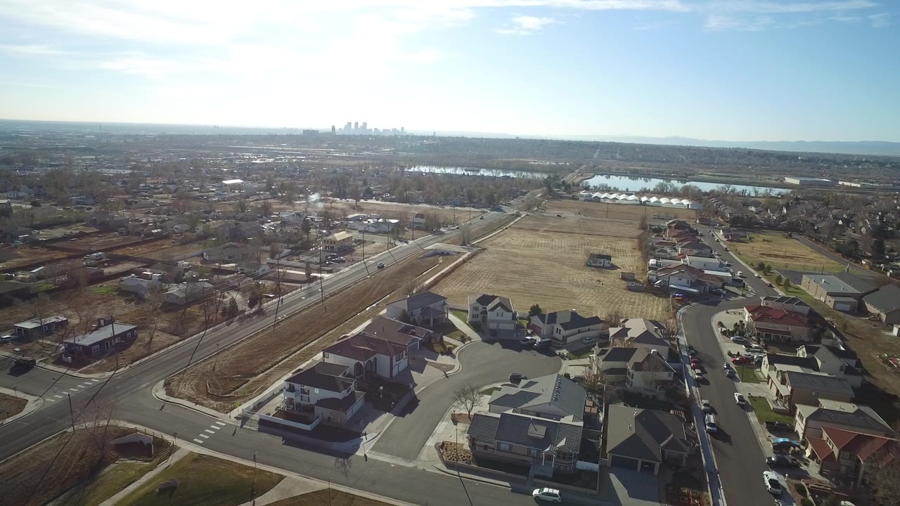 Aerial frame over Westminster, Colorado, with a road intersection and houses below, dry open land to the right, reflective water in the distance, and no visible people.